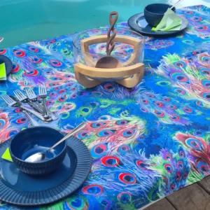 A vibrant table setting by a pool, featuring a colorful tablecloth with a peacock feather design. The table is set with dark blue dishes, silver cutlery, and a clear centerpiece with a wooden decorative item inside. The peacock feather pattern on the tablecloth showcases bright blues, greens, and reds, adding a lively and exotic touch to the outdoor dining experience.