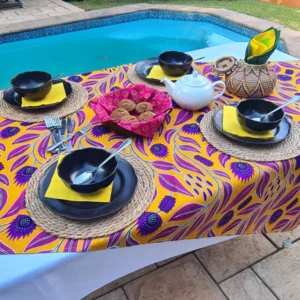 An outdoor dining setup by the pool featuring a vibrant handmade Africa-inspired tablecloth with an exotic floral design in purple and magenta on a golden background, paired with black dishware, woven placemats, and a teapot, creating a warm and inviting atmosphere.