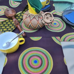 Handcrafted reversible table runner and placemats set featuring bold medallion Shweshwe design with vibrant yellow and green geometric patterns, paired with wooden utensils and colorful tableware on a dining table.