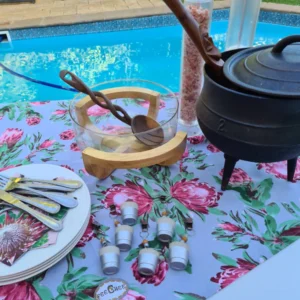 Handmade tablecloth with protea flowers print, adorned with 6 silver-dipped weights, set up on an outdoor table by a pool, surrounded by greenery, creating an elegant and secure outdoor dining setup.